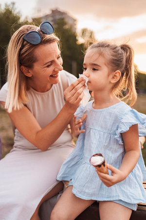 Mother taking care of her daughter who is eating ice cream in a park at sunsetの写真素材