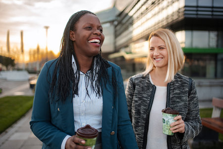 Two businesswomen laughing while walking and holding coffee cups outdoorsの写真素材
