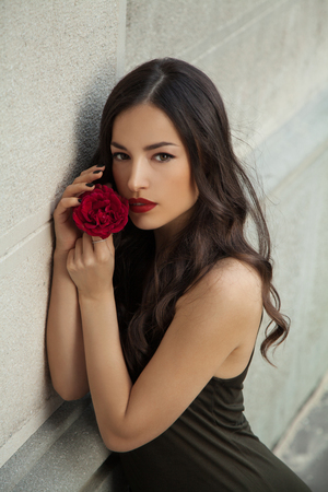 beautiful young woman holding rose close to her face leaned on a white wall, outdoor portraitの写真素材