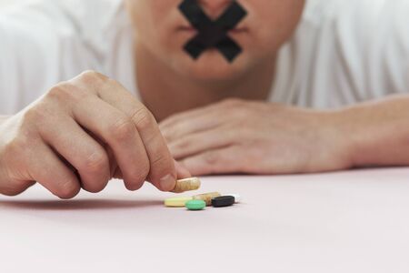 pills on the table, man holds tablet with sticky tape over his mouthの写真素材