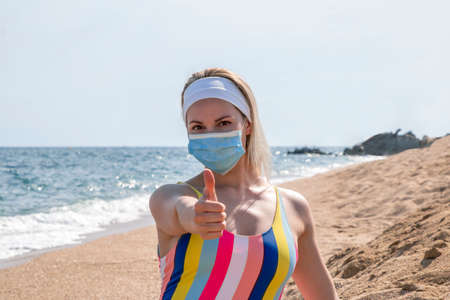 a beautiful girl in a medical mask on the beach shows thumbs up