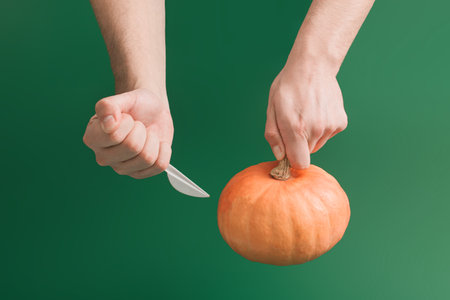 Creative Halloween concept with manâs hands holding pumpkin and knife. Dark green moody background. Aesthetic spooky decoration idea.の写真素材