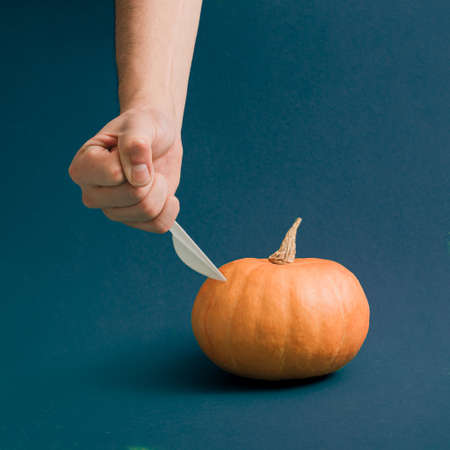 Aesthetic Halloween and autumn idea with a manâs hand holding a knife on a pumpkin. Cyan moody background. Creative season and craft concept, visual minimal arrangement.の写真素材