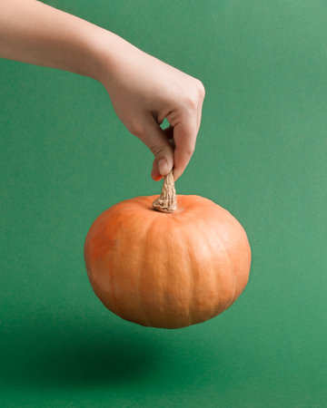 Minimal autumn and Halloween concept with a female hand holding a fresh pumpkin against moody green background. Aesthetic retro graphic idea, vertical simple composition.の写真素材
