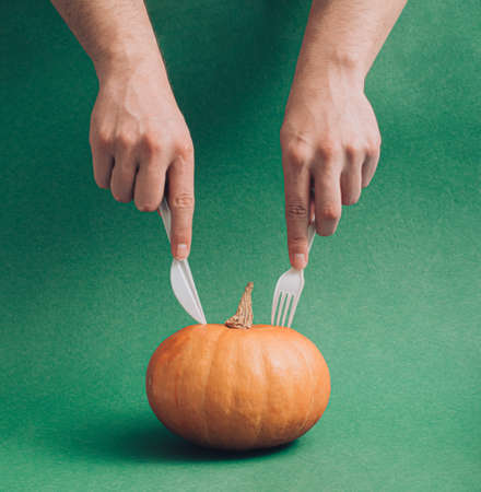 Minimal autumn and Halloween idea with male hands holding fork and knife against fresh pumpkin. Creative holiday season concept, aesthetic simple composition. Moody dark green background.の写真素材