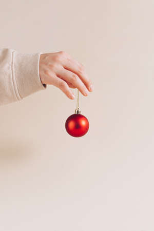 Minimal Christmas and New Year concept with female hand holding red bauble. Pastel beige background, creative season dÃ©cor idea. Visual vertical arrangement.の写真素材