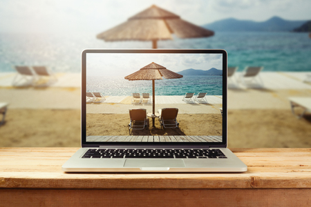 Laptop computer with sunny beach image on wooden table. Summer vacation photo sharingの写真素材