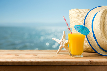Summer holiday vacation concept with orange juice, hat and bag over sea beach backgroundの写真素材