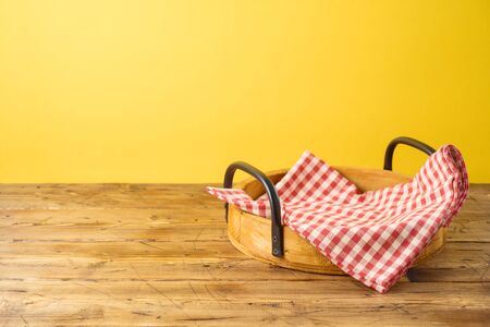 Empty wooden tray with red checked tablecloth on wooden table over yellow background