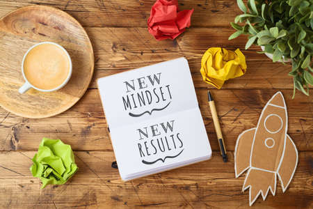 New mindset and new results text on notebook on wooden table with coffee cup and paper rocket. Business concept of positive thinking and motivationの写真素材