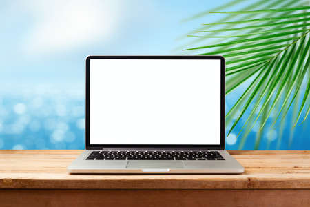 Laptop computer with blank screen on wooden table over sunny tropical beach background. Summer vacation mock up for designの写真素材