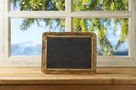 Chalkboard on wooden table over window with winter landscape background.  Christmas and New Year mock up for design and product display.の写真素材