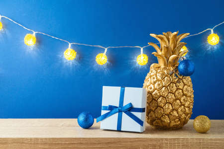 Christmas holiday greeting card with pineapple as alternative Christmas tree, lights garland and gift box on wooden shelf over blue backgroundの写真素材