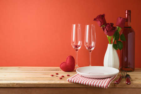 Valentines day romantic dinner concept. Wooden table with plate, wine bottle and rose flowers over red backgroundの写真素材