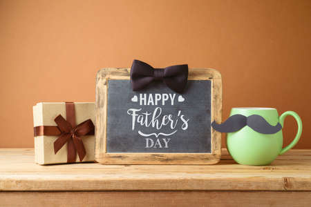 Happy Fathers day greeting card with green coffee cup, mustache, gift box and chalkboard on wooden tableの写真素材