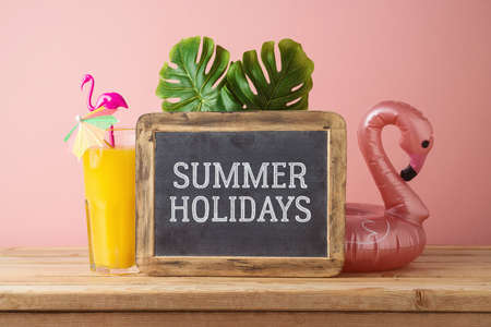 Summer holidays concept with orange juice, flamingo pool float and chalkboard on wooden table over...