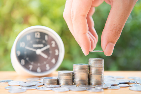 Human hand is putting coin to growing coin stack with clock and nature background for saving money...