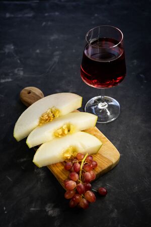 selective focus, clear glass of red grape wine. Olive wood Board with cut melon and pink grapes. Still life, low key, with copyspaceの写真素材