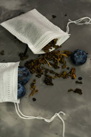preparation for tea. craft tea bags with blueberry berries and green leafy Oolong tea on a grey textured background. For brewing in a mugの写真素材