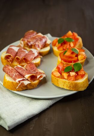 selective focus, snack bruschetta, with red tomatoes and Basil, and smoked meats. On a small white bread baguette on a dark backgroundの写真素材