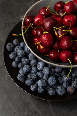 Selective focus, ripe cherries and blue blueberries with water drops in a dark kitchen dish, on a dark background, vertical position, rustic styleの写真素材