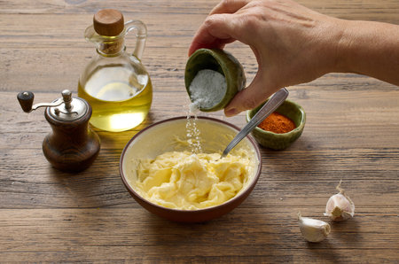 adding salt to bowl of whipped butter. garlic butter ingredients on wooden kitchen tableの写真素材