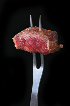 Freshly grilled piece of beef steak meat on a fork isolated on a black background. Close-up of foodの写真素材