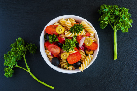 Salad: pasta fusilli, black and green olives, cherry tomatoes, red onion and parsley. Dressing: olive oil and lemon juice. In white bowl. Black stone background. Top view.の写真素材