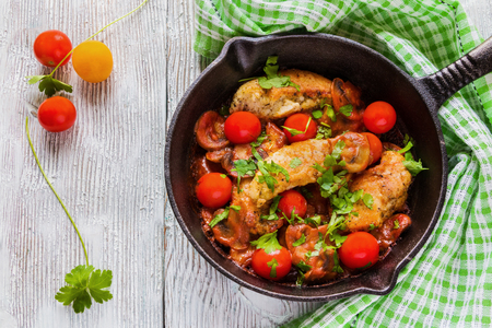 Roasted chicken fillet, cooked with mushrooms, garlic, paprika and olive oil. Cast-iron skillet and fresh cherry tomatoes on wooden table, top view.の写真素材