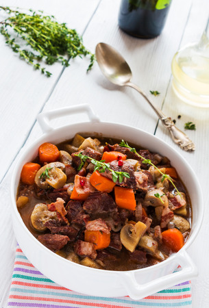 Beef Bourguignon in a casserole on white table. Stewed with bacon, garlic, carrots, onions, mushrooms,  and spices. Bunch of fresh thyme, vintage spoon, red wine.の写真素材