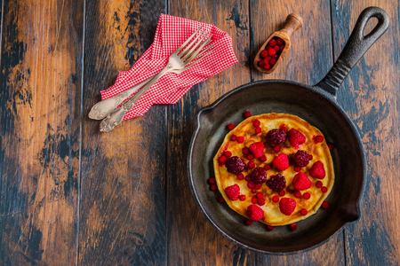 Homemade fried pancakes on a black cast iron skillet. Above are berries, raspberries, cranberries and blackberries. Wooden rustic brown background, top view.の写真素材