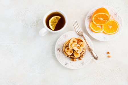 Stack of homemade pancakes with banana, maple syrup and walnuts on vintage plate. Fork, fresh sliced fresh lemon, cup of tea, white and gray concrete background, top view.の写真素材