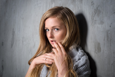 Sexy young longhair blonde woman in knitted sweater posing against grungy gray wall. Scared or resentful attractive girl hiding behind a lock of her hair.の写真素材