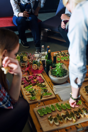 Company of friends eating together in the restaurant. Wooden tray of appetizers at a banquet on a decorated pallet coffee table and carafes of fruity beverage with lemon and mintの写真素材