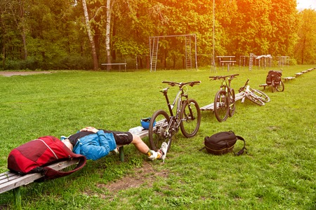 Cyclist resting on a bench in the park. Tired young man with his mountain bike in the forest resting and making pauseの写真素材