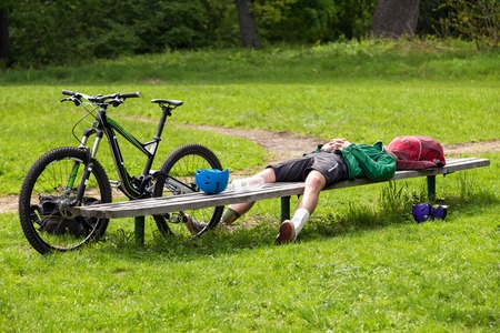 Cyclist resting on a bench in the park. Tired young man with his mountain bike in the forest resting and making pauseの写真素材