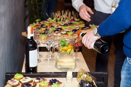 Waiter pouring red wine in a glass at a restaurant table full of appetizers with guests standing nearの写真素材
