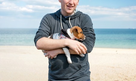 Young happy man holding his cute small dog Jack Russell terrier on beachの写真素材