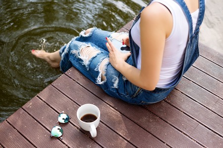 young woman using her smartphone sitting on the pier and drinking hot coffee.の写真素材