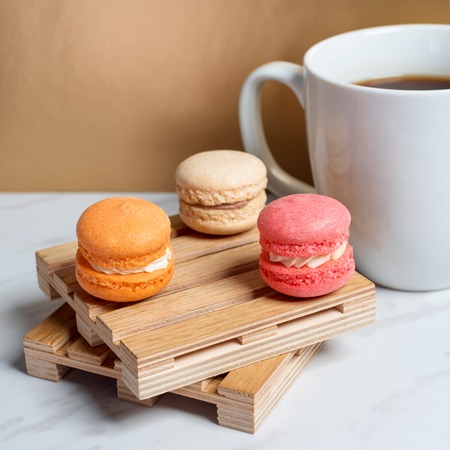 Sweet and colourful macaroons served on little wooden pallets with cup of coffee on a marble texture table and golden background. Traditional french dessert. Copy space.の写真素材