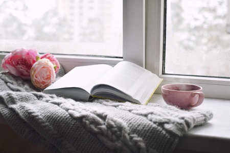 Cozy home still life: cup of hot coffee, spring flowers and opened book with warm plaid on windowsill. Springtime concept, free copy spaceの写真素材
