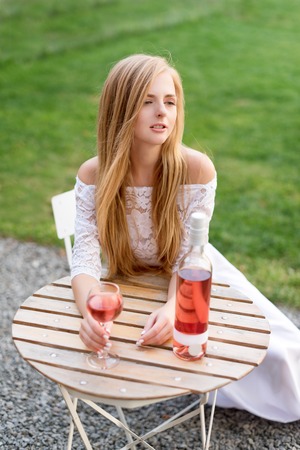 Beautiful woman drinking wine in outdoors cafe. Portrait of young blonde beauty in the vineyards having fun, enjoying a glass of wine in the garden or tasting alcohol cocktail at dining table in lawn.の写真素材