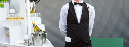 Midsection of professional waiter in uniform serving wine during buffet catering party, festive event or wedding. Full glasses of champagne on tray. Outdoor party catering service, waiter on duty.の写真素材
