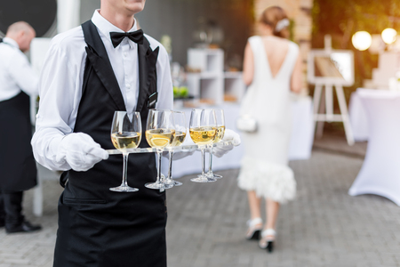 Midsection of professional waiter in uniform serving wine during buffet catering party, festive event or wedding. Full glasses on tray.の写真素材