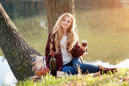 Beautiful woman drinking wine outdoors having picnic in the park. Portrait of young blonde beauty enjoying a glass of red wine sittind on autumn lawn near the lake.の写真素材