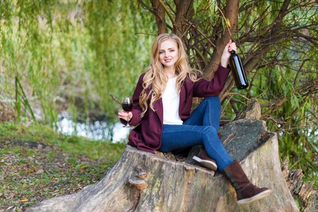 Beautiful woman drinking wine outdoors having picnic in the autumn park. Portrait of young blonde beauty enjoying a glass of red wine sitting on giant tree stump near the lake.の写真素材
