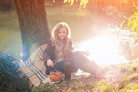 Beautiful woman drinking wine outdoors having picnic in the park. Portrait of young blonde beauty enjoying a glass of red wine sitting on autumn lawn near the lake.の写真素材