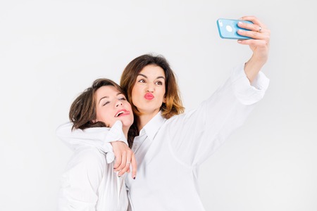 Two women posing taking selfie in light studio. Girls best friends making funny grimaces on camera, showing tongue and laughing together.の写真素材