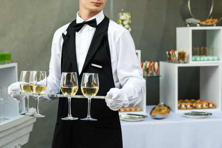 Midsection of professional waiter in uniform serving wine during buffet catering party, festive event or wedding. Full glasses of champagne on tray. Outdoor party catering service, waiter on dutyの写真素材