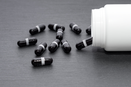 Black supplement capsules are poured from white bottle on slate board. Medicine theme, health care conceptの写真素材